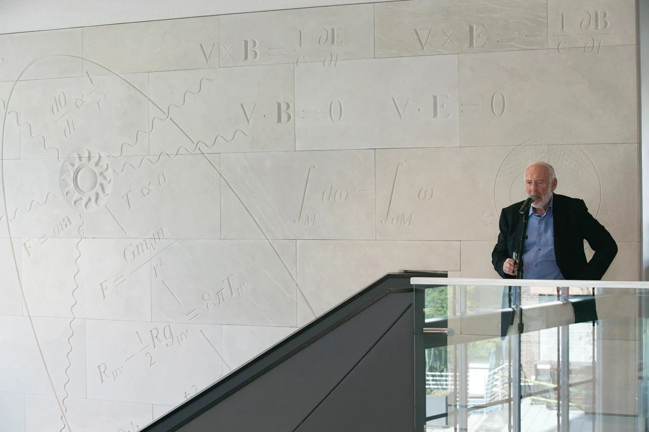 James Simons speaking at a microphone in front of the Iconic Wall